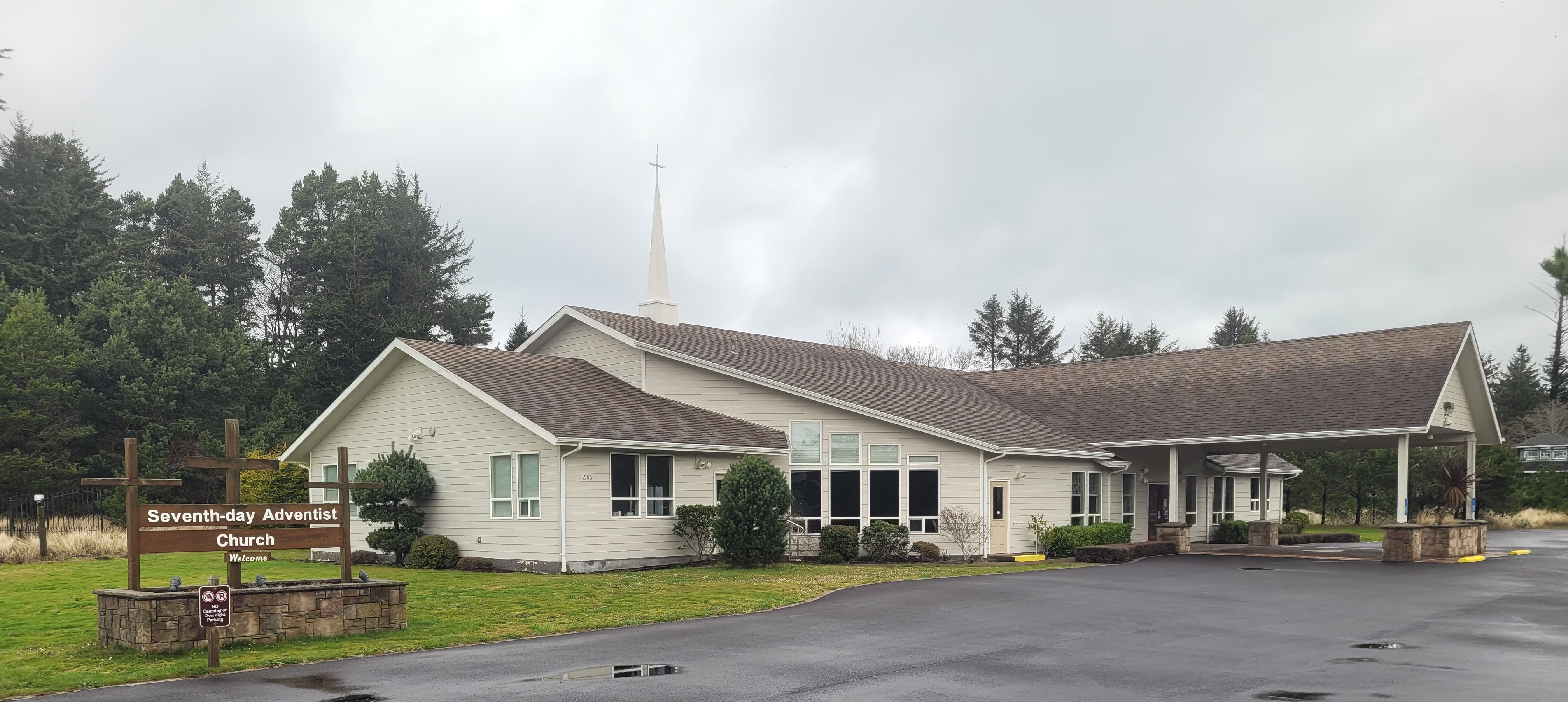 A beige building with a steeple serves as a Seventh-day Adventist church. The sign outside reads "Seventh-day Adventist Church" with a direction arrow. Overcast sky in the background.