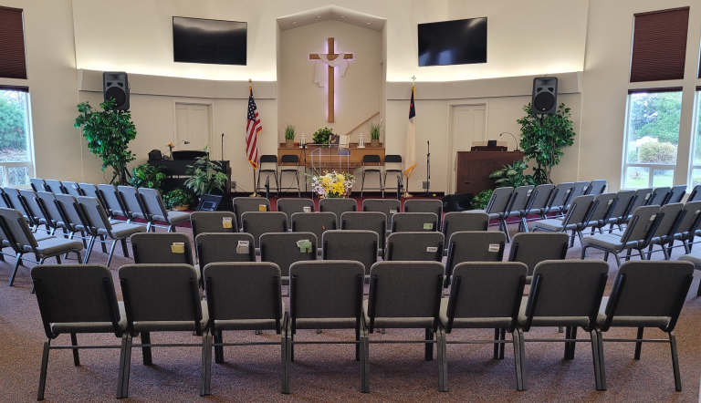 A church auditorium with rows of gray chairs facing a stage with a cross, two screens, flags, plants, and musical equipment.