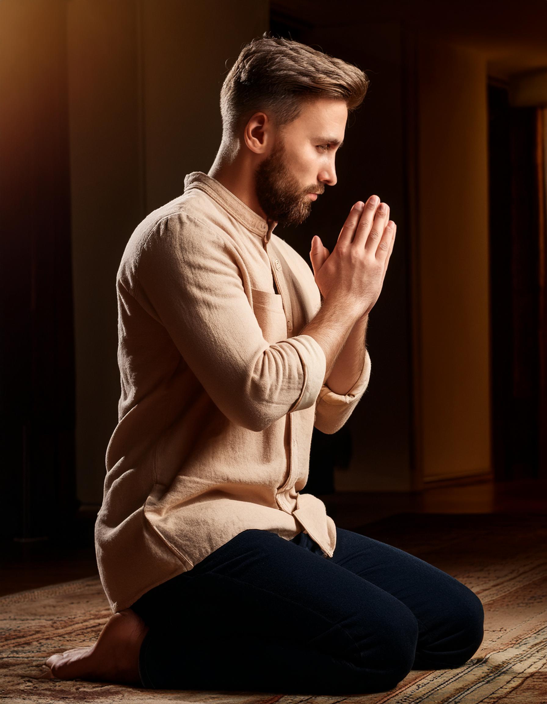A man with a beard kneels on a rug indoors, hands pressed together in front of his face in a prayer or meditation pose.