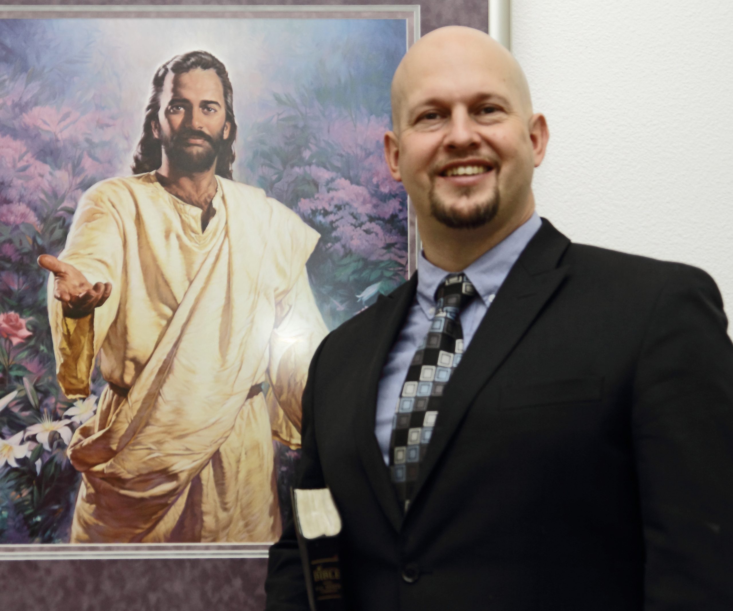 A man in a suit holding a Bible stands in front of a framed painting depicting Jesus with an outstretched hand.